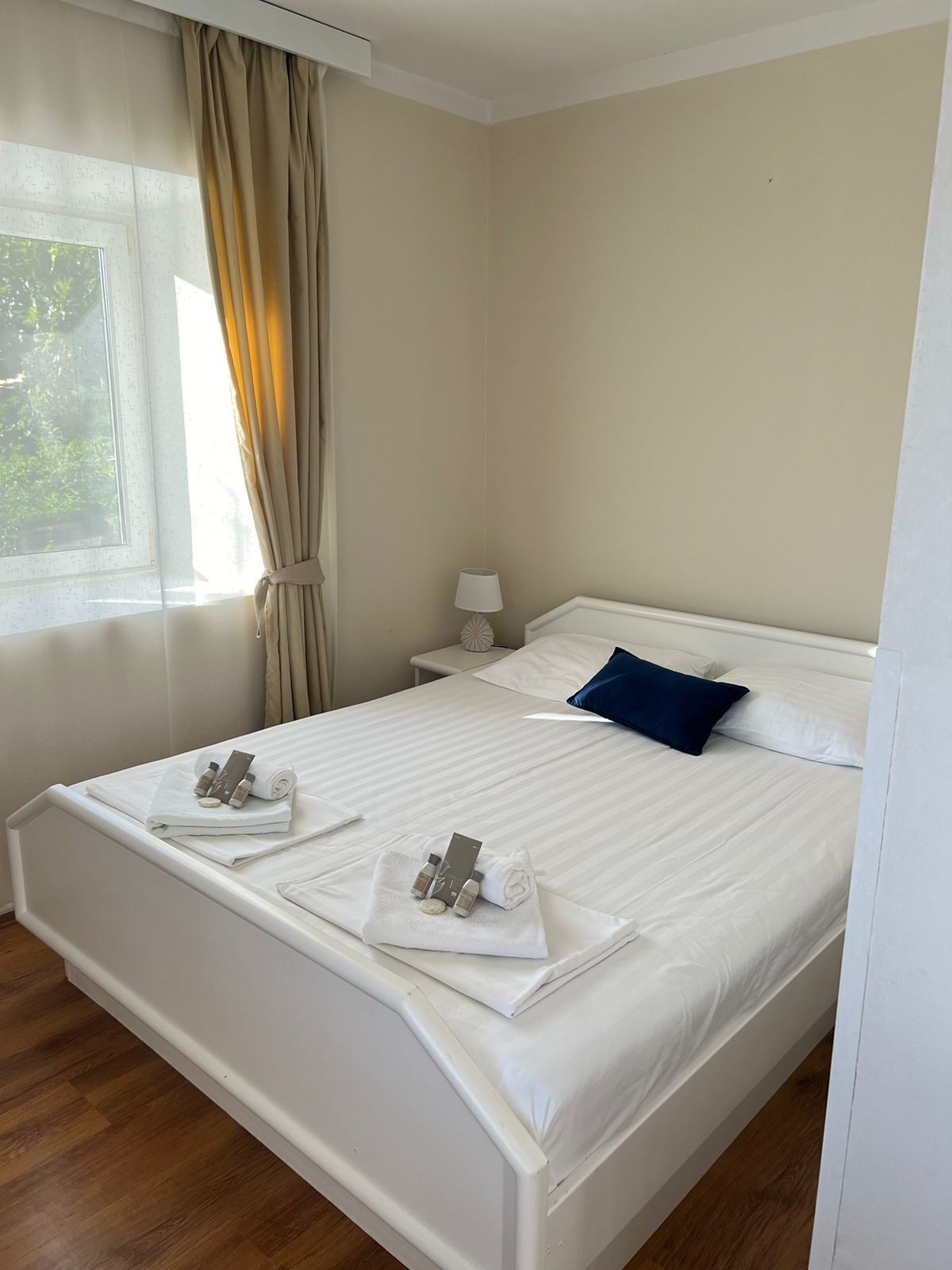 A neatly made bed with towels and toiletries, a bedside lamp, and a window with curtains.