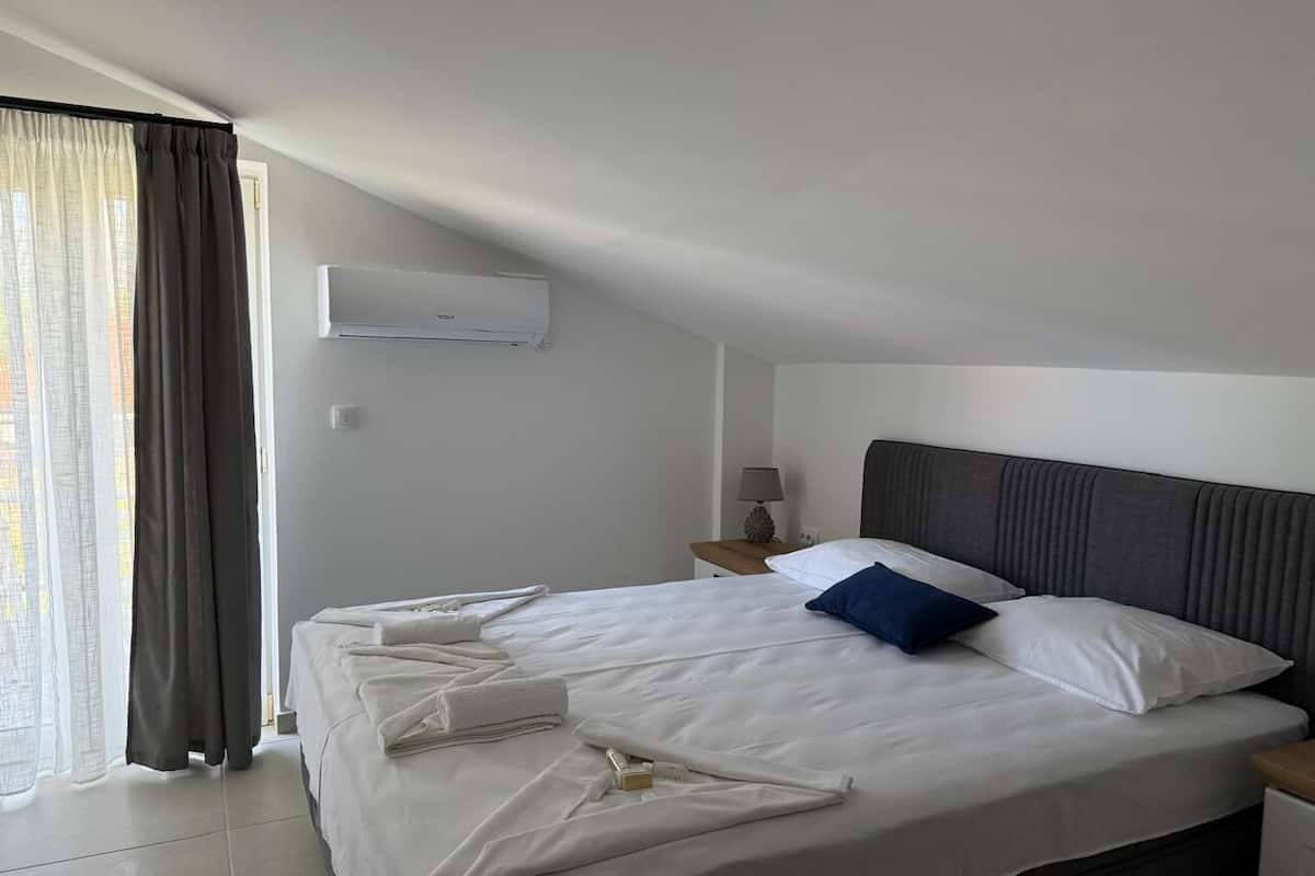 Una cama bien hecha con sábanas blancas y almohada azul, una mesita de noche con lámpara, y una ventana con cortinas.
