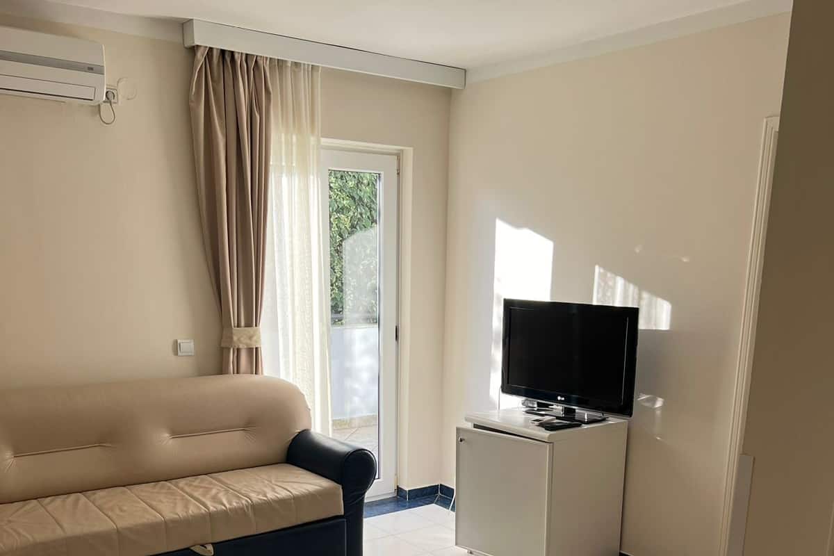 Una sala de estar con un sofá color beige, un televisor sobre un mueble blanco y un ventanal corredizo que da a un espacio exterior.