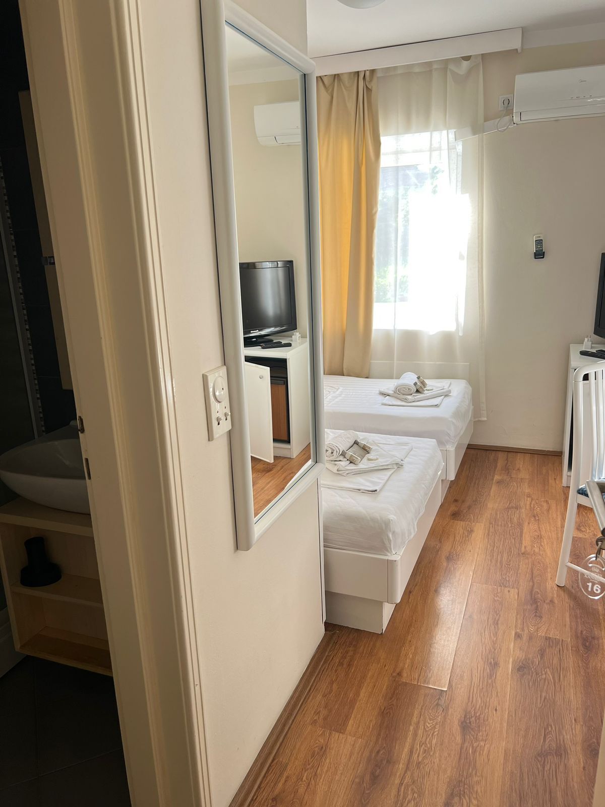 Habitación de hotel con piso de madera, una cama, un televisor y un baño con lavamanos y espejo.