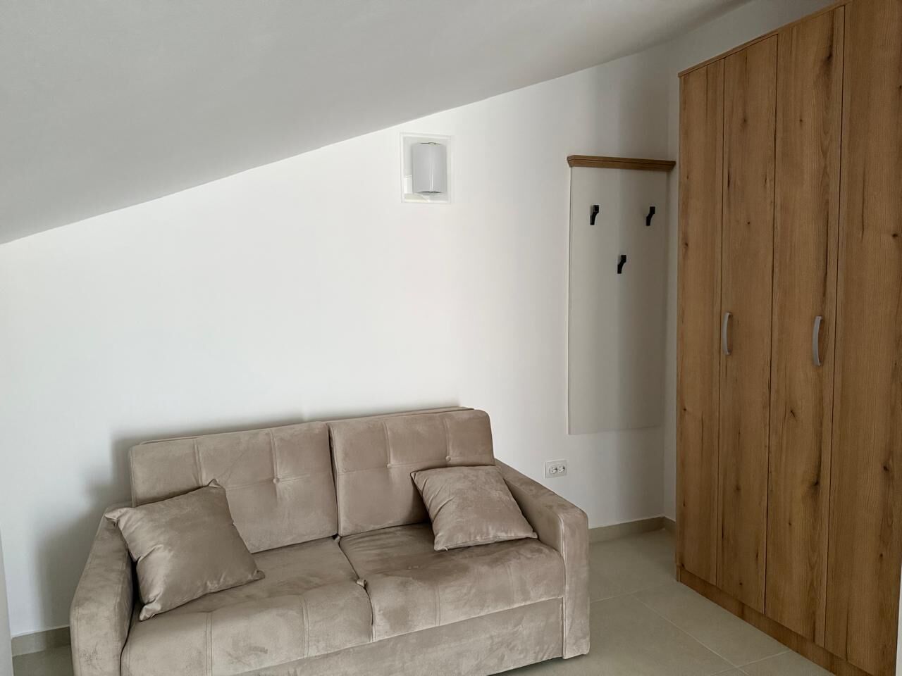 A room with a beige sofa, a wooden wardrobe, and a white wall with a small light fixture.