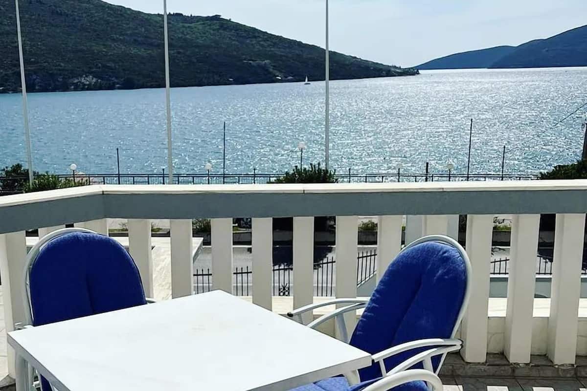 Una terraza con una mesa blanca y sillas azules, con vista a un lago tranquilo y colinas al fondo.