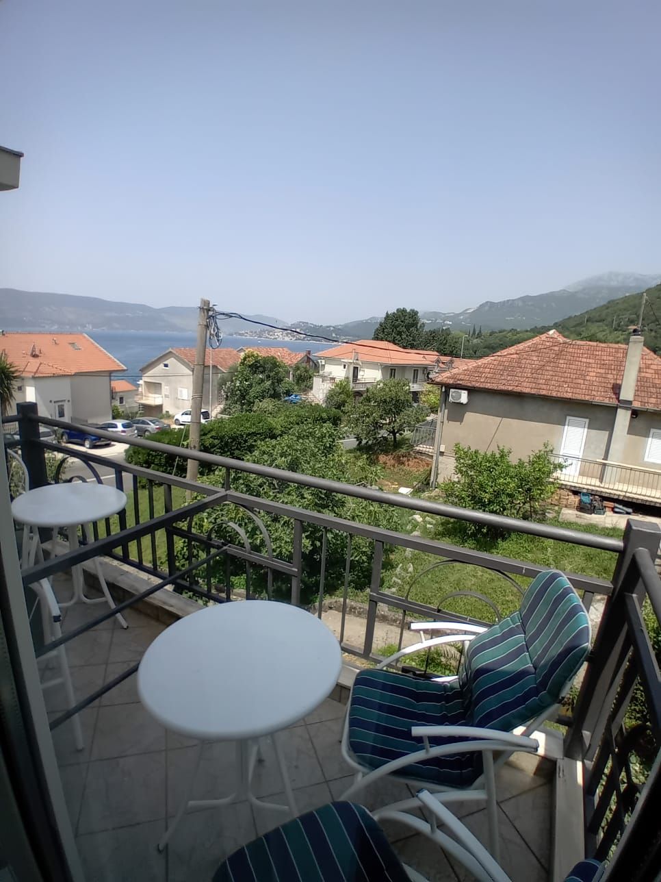 Un balcon avec une table blanche et des chaises à rayures, donnant sur un quartier résidentiel avec des maisons et offrant une vue lointaine sur la mer.