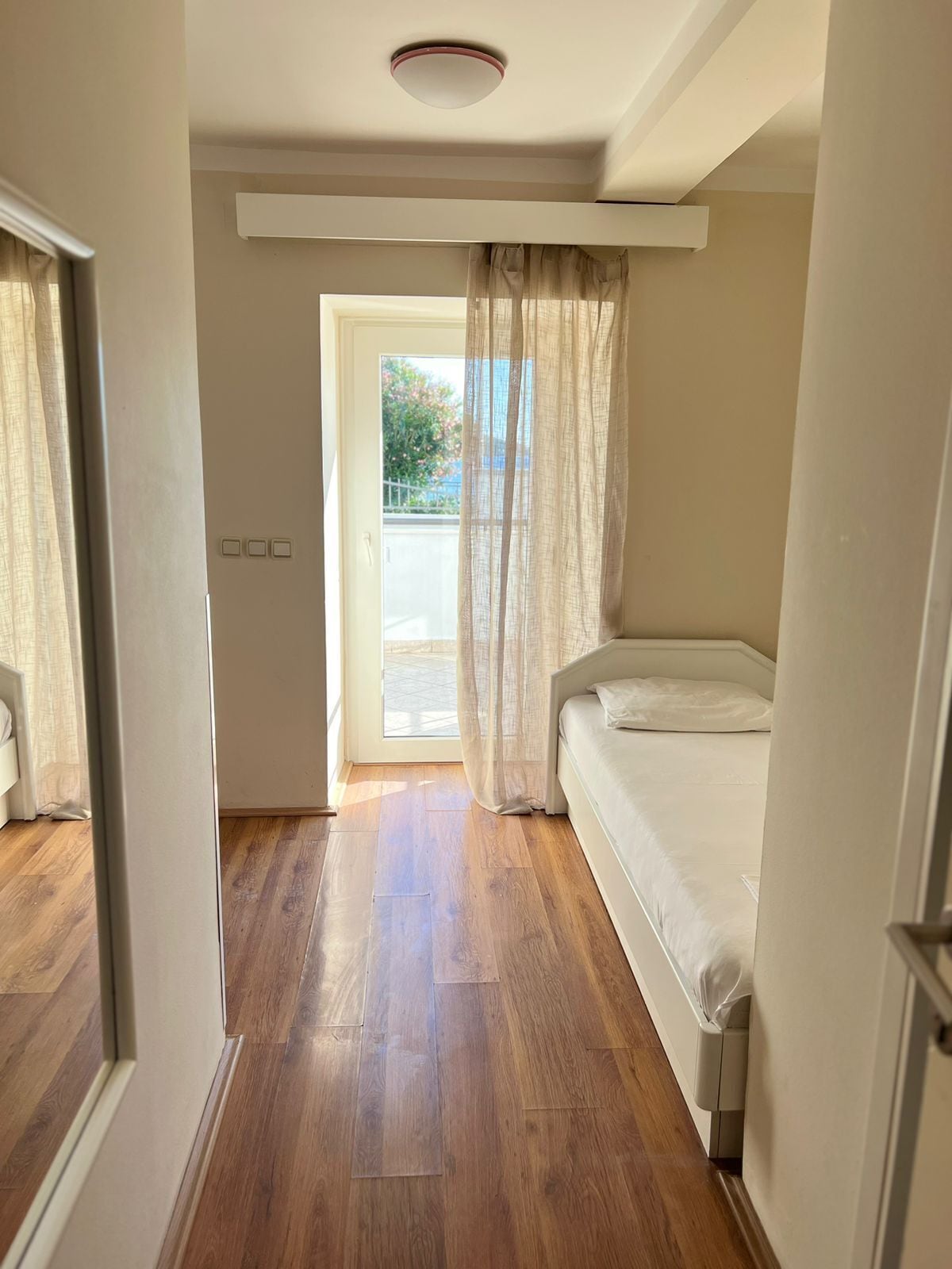 Un dormitorio con piso de madera, una cama, un ventanal con cortinas de gasa y vistas al exterior.