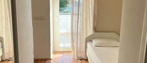 Un dormitorio con piso de madera, una cama, un ventanal con cortinas de gasa y vistas al exterior.