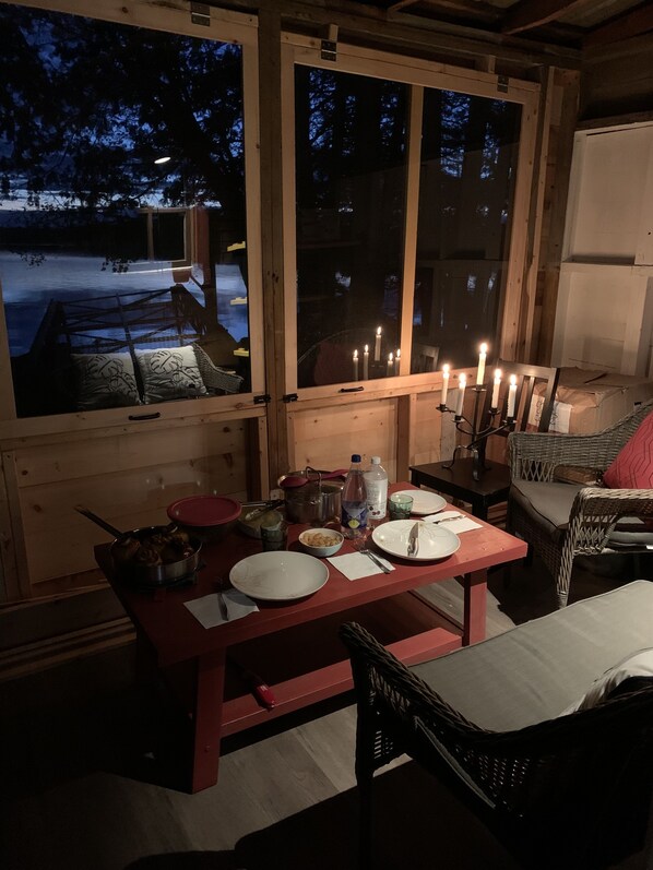 L’intérieur chaleureux d’un chalet avec une table à manger dressée pour deux personnes, un fauteuil en osier et une vue sur un paysage enneigé par de grandes fenêtres.