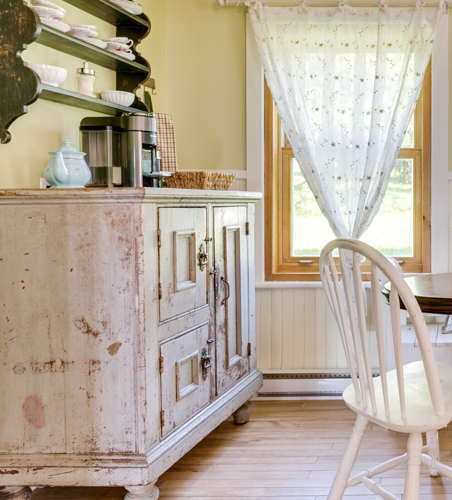 Eine rustikale Küche mit einem weißen Schrank, einem Holzstuhl und einem Fenster mit floralem Vorhang.