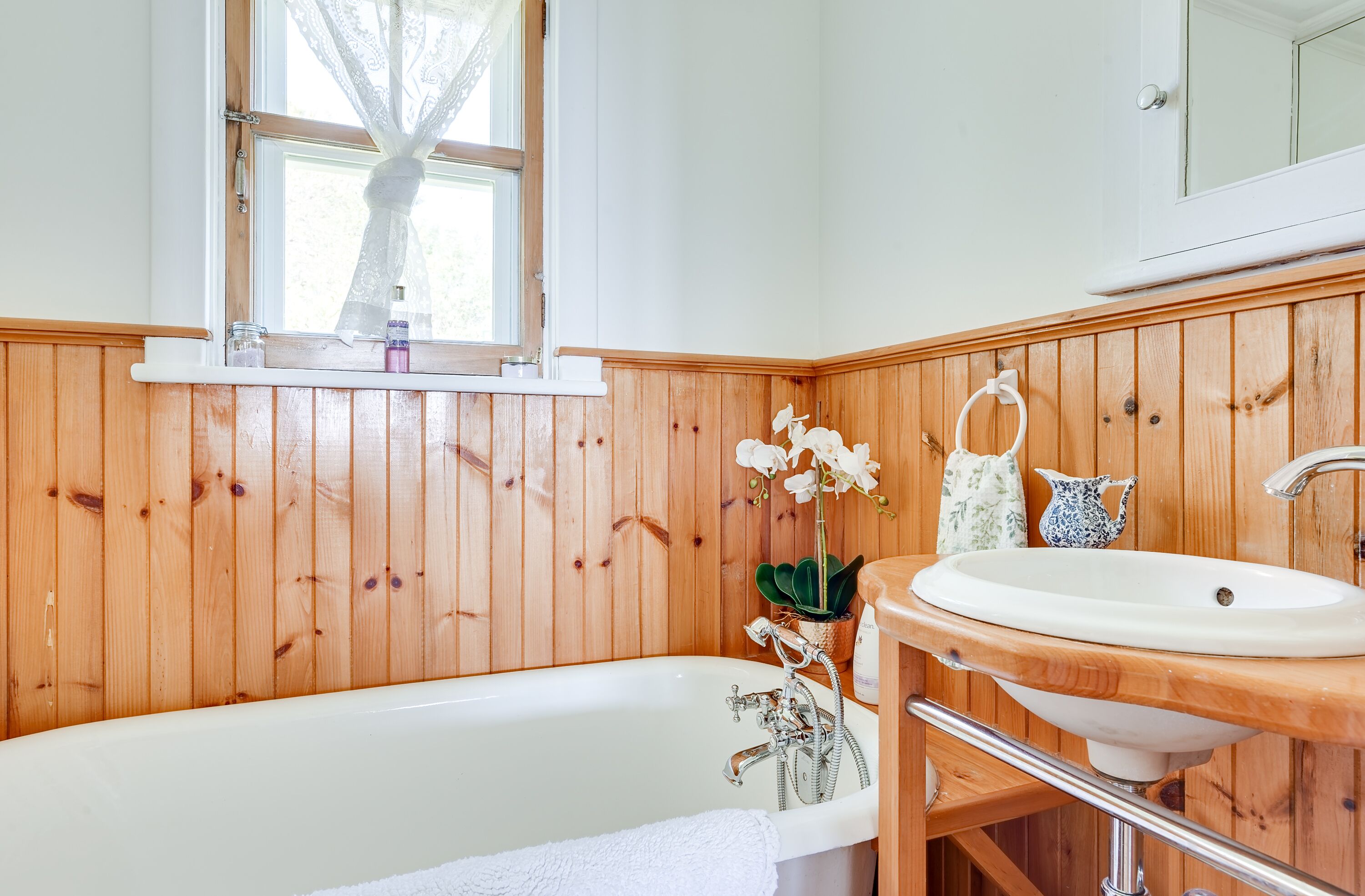 A bathroom with a wooden panel wall, a white bathtub, a wooden vanity with a sink, and a window with a lace curtain.