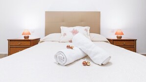 Ein ordentlich bezogenes Bett mit weißer Daunendecke, zwei Kissen mit Blumenmuster und ein aufgerolltes weißes Handtuch. An beiden Seiten des Bettes befinden sich jeweils zwei Nachttische mit Lampen.