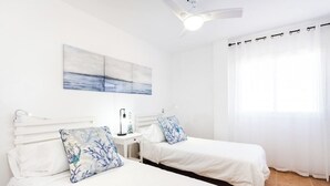 A hotel room with two beds, a nightstand, a lamp, a ceiling fan, a window with curtains, and a painting on the wall. - Home2Book Stunning Apartment Candelaria, Terrace (Las Caletillas)
