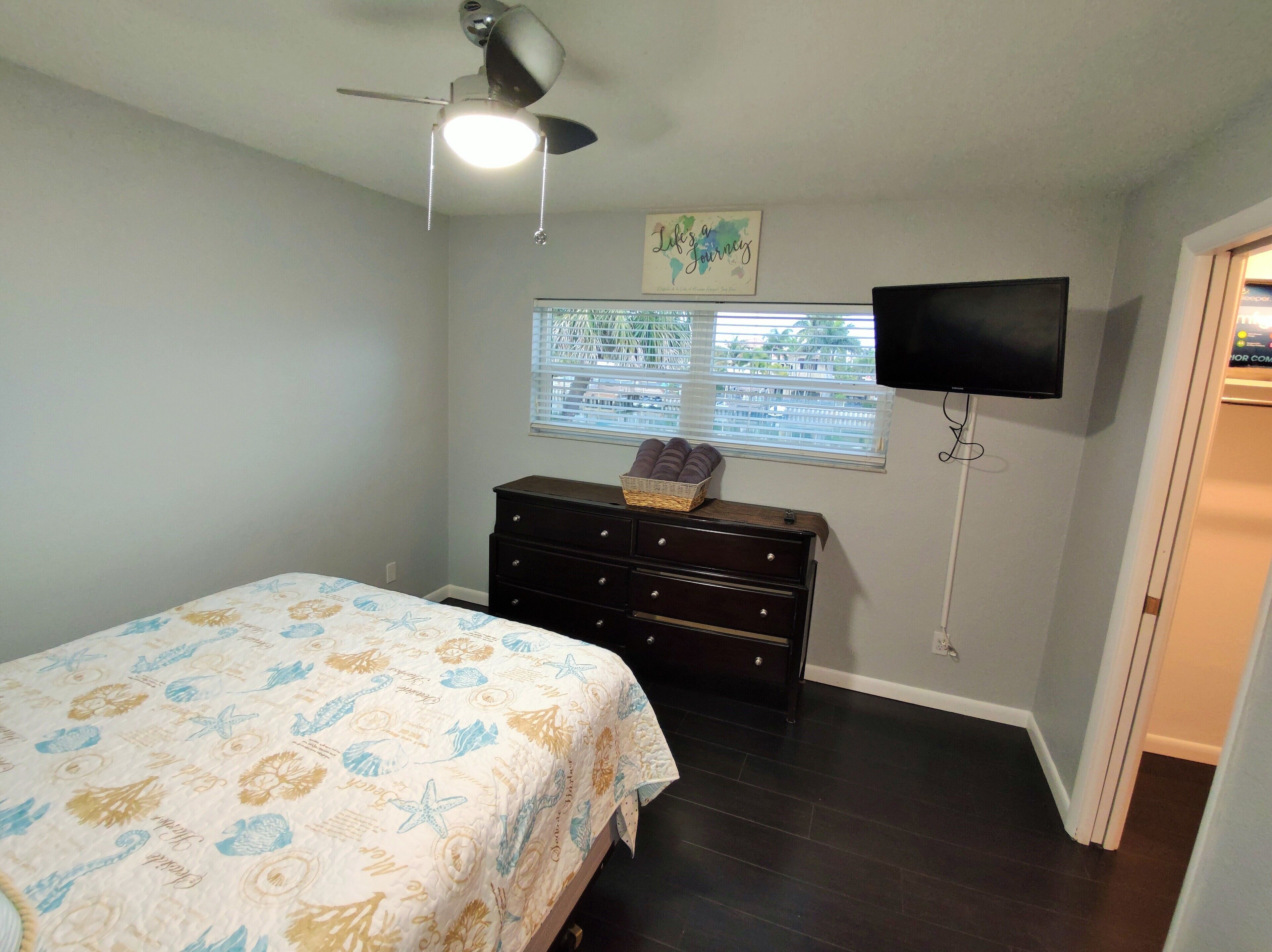 A bedroom with a bed, a dresser, a ceiling fan, a window with blinds, and a TV mounted on the wall.