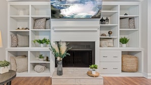 A living room with a fireplace, shelves with decorative items, and a television displaying a waterfall scene. - Hamilton Hideaway- Pool, Creek View w/ Scenic Deck (Mobile)