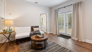 A modern living room with a white sofa, a round wooden coffee table, and a small side table with a lamp and bottles. - Hamilton Hideaway- Pool, Creek View w/ Scenic Deck (Mobile)