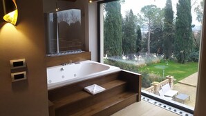 A modern bathroom with a large bathtub, wooden steps, and a view of a garden through a large window. - Le Cappuccinelle Suites&SPA (Perugia)