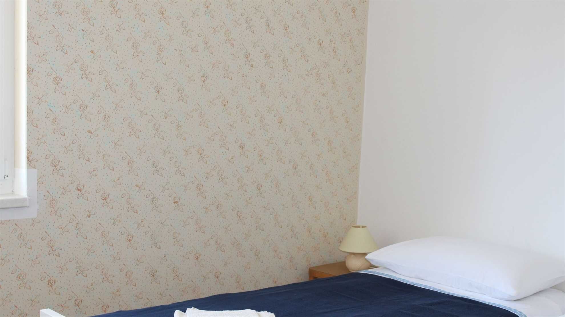 Una camera da letto con carta da parati a motivi floreali, un letto con lenzuola bianche e azzurre, una lampada da comodino e una finestra.
