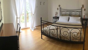 Ein Schlafzimmer mit einem gebogenen Metallbettgestell, Holzboden, einem kleinen Nachttisch mit Lampe und einem Sessel.