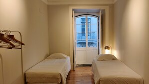 A narrow hotel room with two single beds, a wooden floor, a coat rack with hangers, and a view through a door to an outdoor balcony. - Dear Home ★ In the Heart of Lisbon (Lisboa)