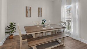 A dining area with a wooden table, benches, and chairs. There are two wall-mounted artworks and a window with white curtains.