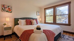 Una cama bien hecha con una colcha roja, una mesita de noche con una lámpara y una ventana con vistas a árboles.