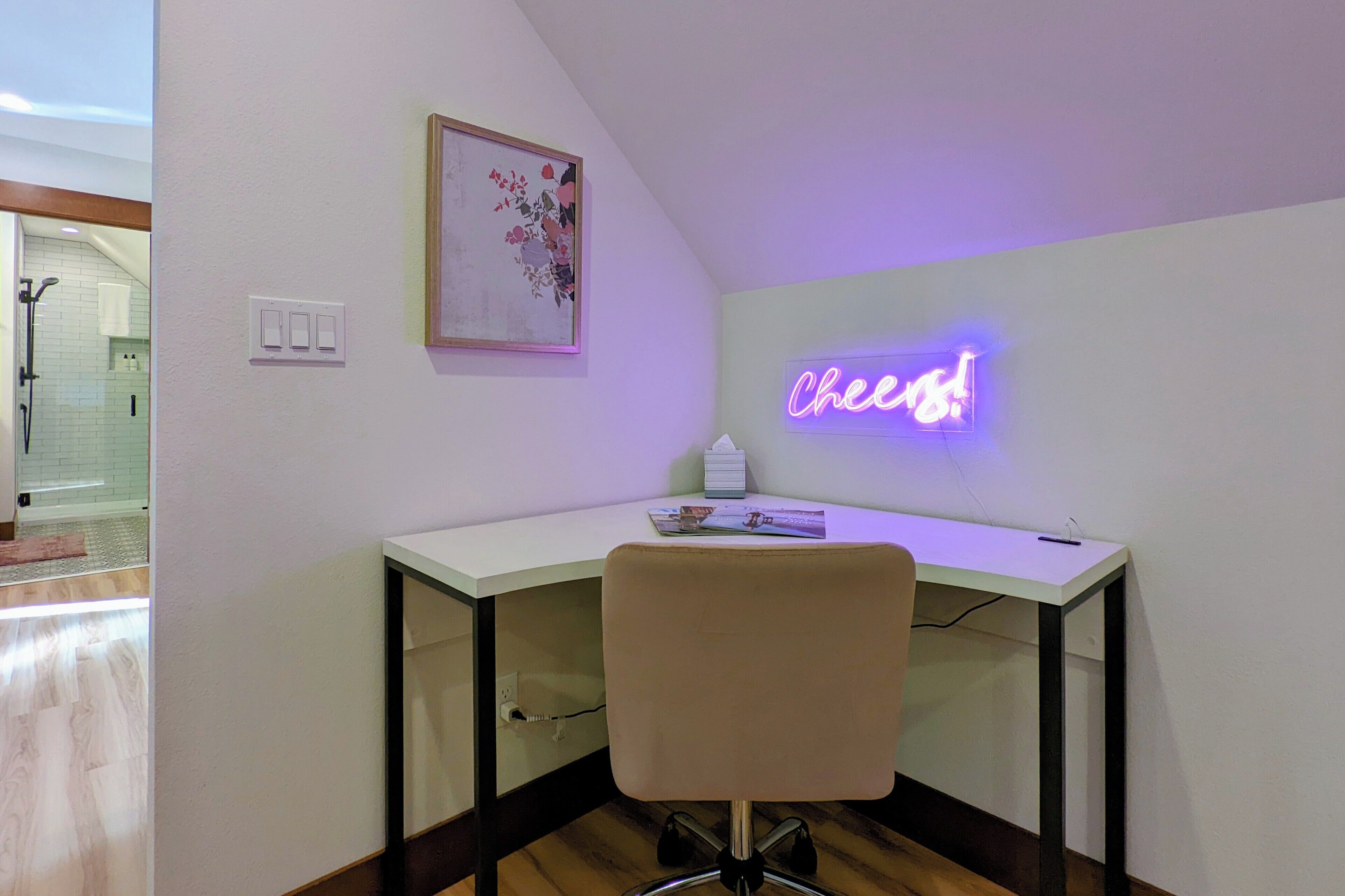 A modern office space with a white desk, a neon sign reading "Cheers!", a framed floral artwork, and a chair.