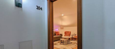A hallway with a door marked '204' leading to a living area with a sofa, armchair, coffee table, and a small table.