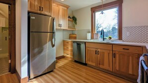 Una cocina con armarios de madera, un refrigerador de acero inoxidable, un lavavajillas y una ventana con vista a árboles.