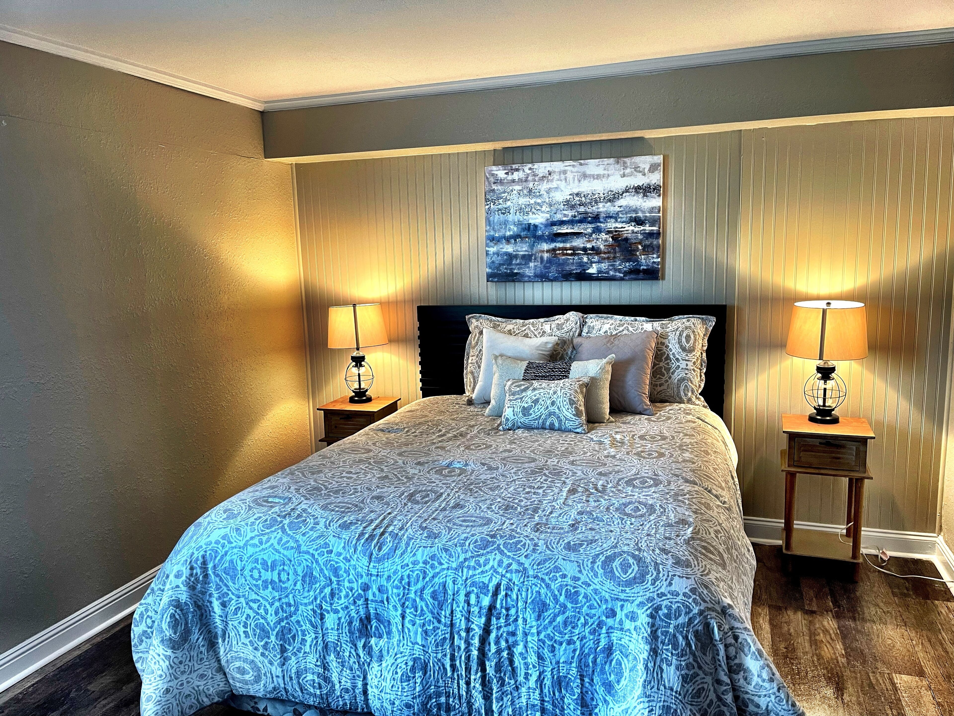 A neatly made bed with patterned bedding, two bedside lamps, and a painting above the headboard.