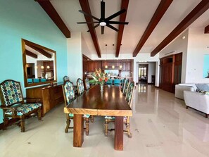 Un'ampia sala da pranzo con un lungo tavolo di legno e sedie decorate, una ventola a soffitto e una cucina sullo sfondo.