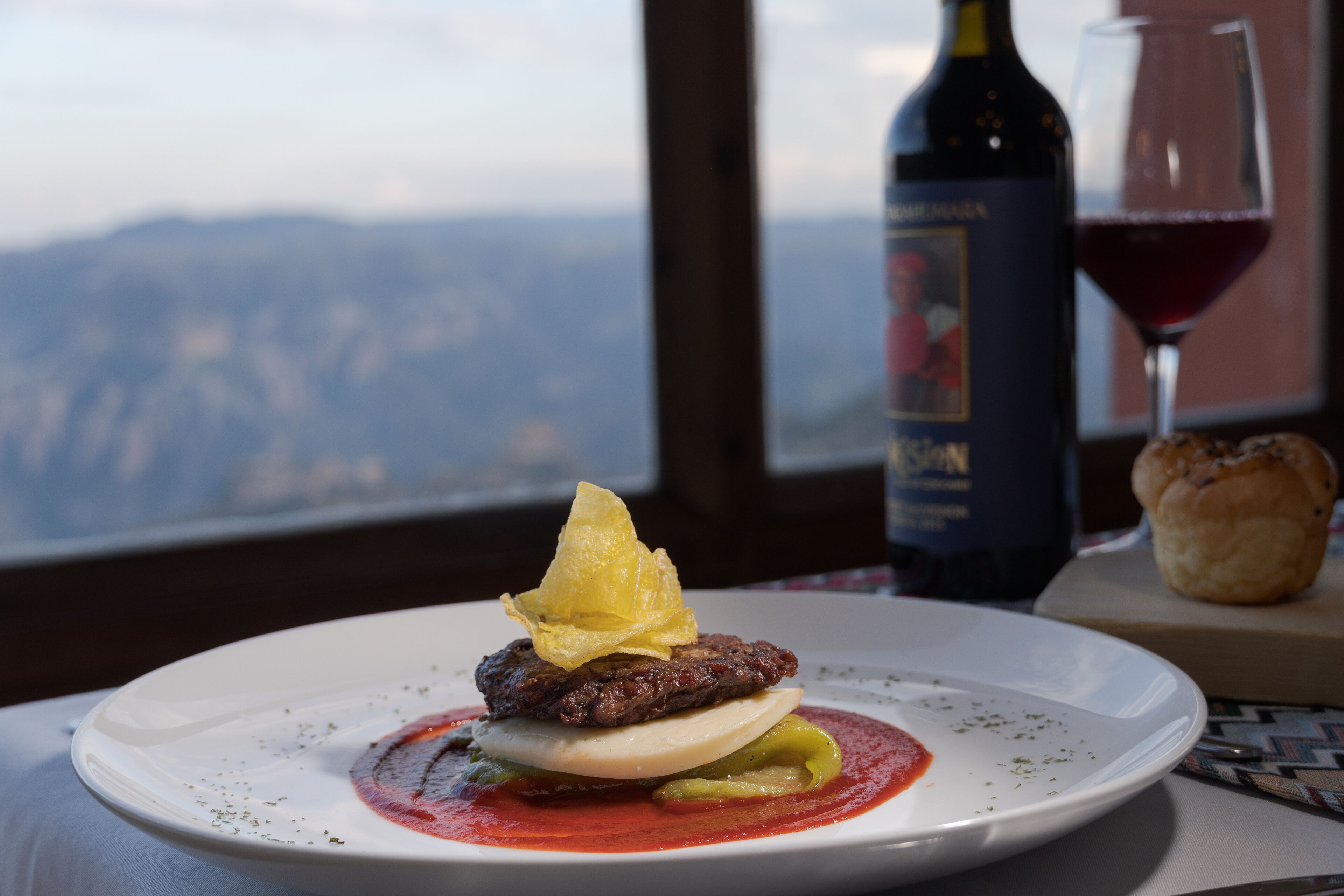 Prato de comida com hambúrguer, ovo e molho, uma garrafa de vinho e um copo de vinho sobre uma mesa com vista para as montanhas ao fundo.