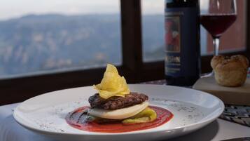 Prato de comida com hambúrguer, ovo e molho, uma garrafa de vinho e um copo de vinho sobre uma mesa com vista para as montanhas ao fundo.