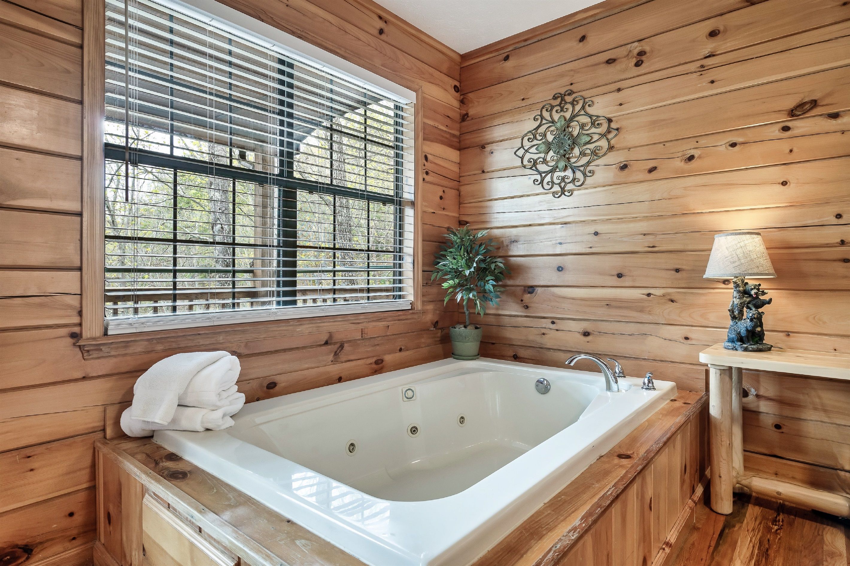 Une salle de bain rustique, avec des murs en bois, comprenant une grande baignoire, une fenêtre avec des stores, une plante en pot et un luminaire fixé au mur.