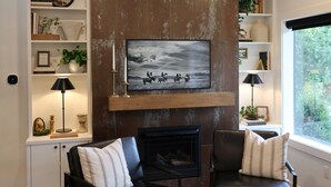 A living room with a fireplace, two leather chairs, a bookshelf, and a large window.