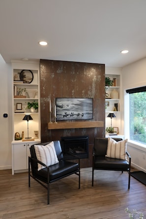 A living room with a fireplace, two leather chairs, a bookshelf, and a large window.