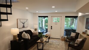 A living room with a black sofa, a coffee table, a lamp, and a painting of an elk. There is a door leading outside and a television on the wall.