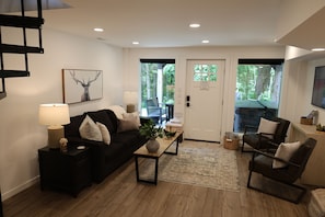 A living room with a black sofa, a coffee table, a lamp, and a painting of an elk. There is a door leading outside and a television on the wall.