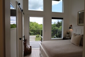 A bedroom with a large bed, a view of greenery through a balcony door, and a small table with a vase.