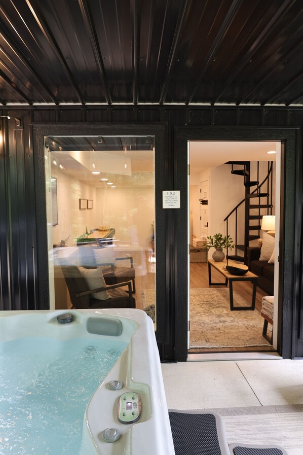 A hot tub with a control panel, a glass door leading to a room with a bed, a staircase, and a living area with a sofa and a lamp.