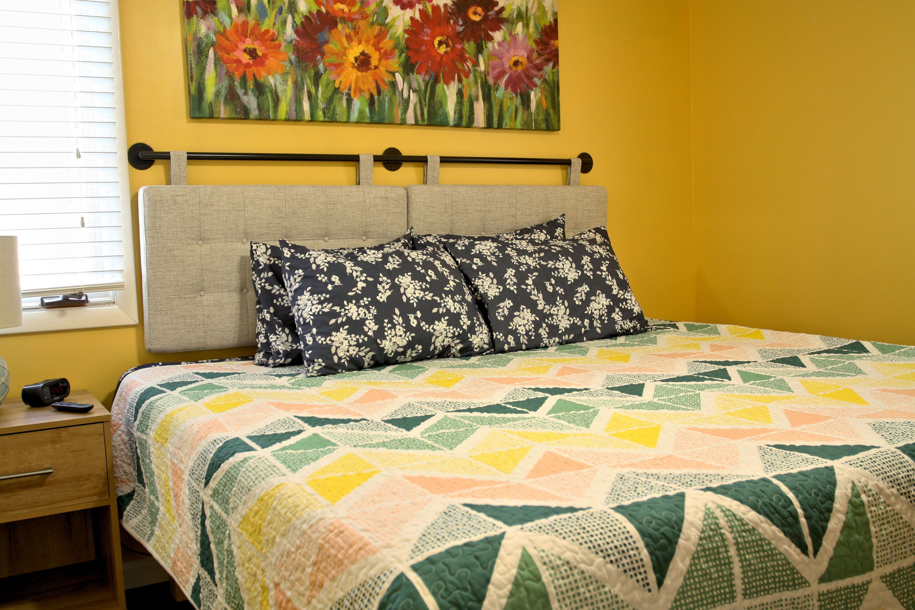 Uma cama com colcha, mesa de cabeceira, quadro e janela com persianas.