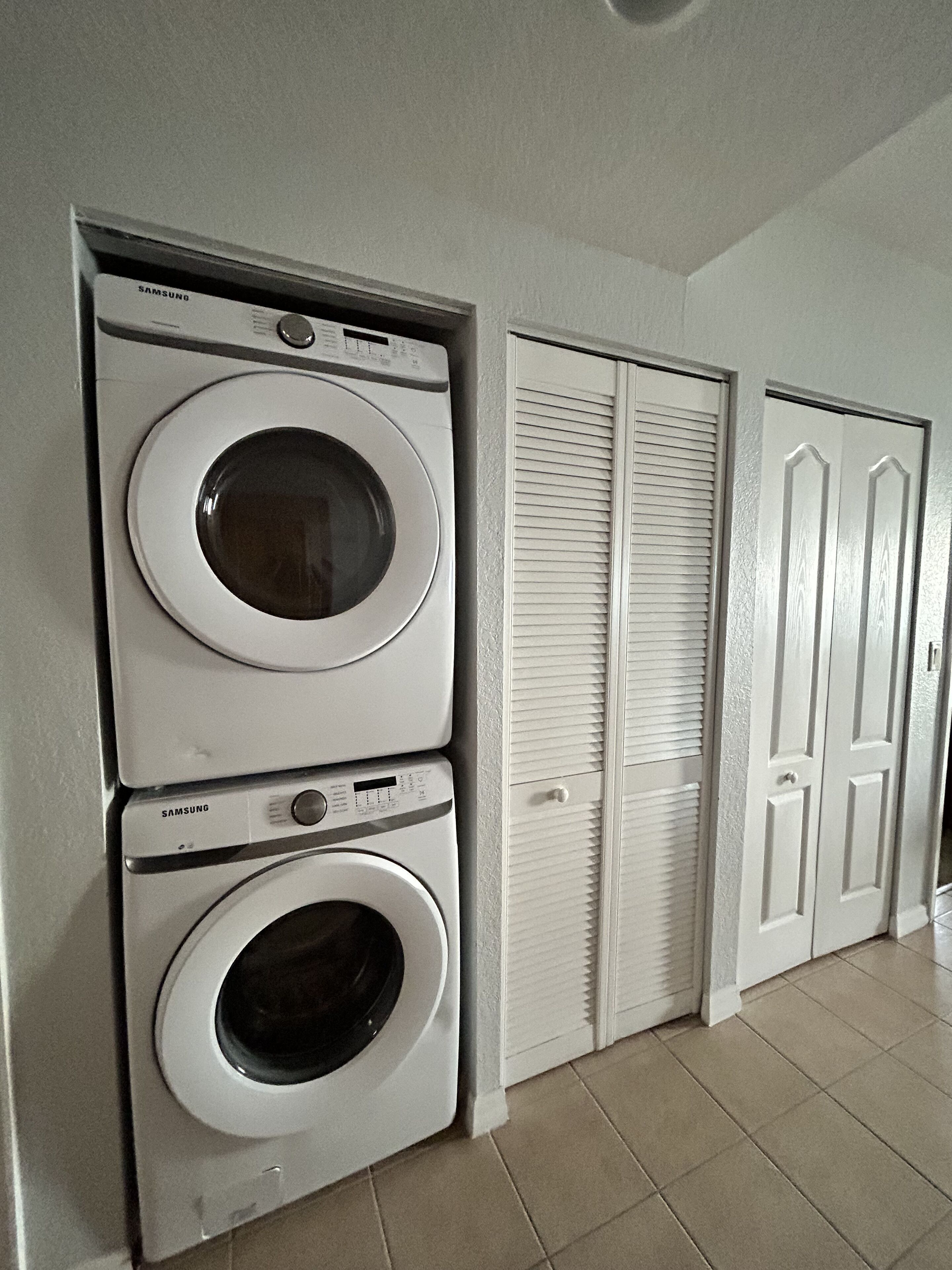 A laundry room with two stacked Samsung washing machines, a white door with louvered shutters, and a tiled floor.