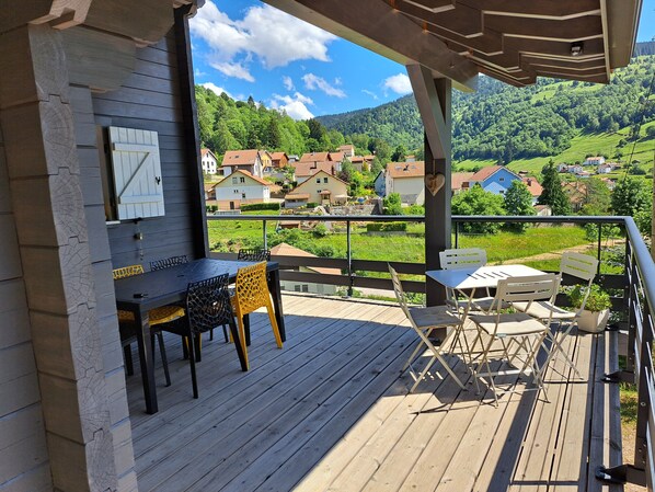 A wooden deck with a table and chairs, overlooking a village with houses and green hills.