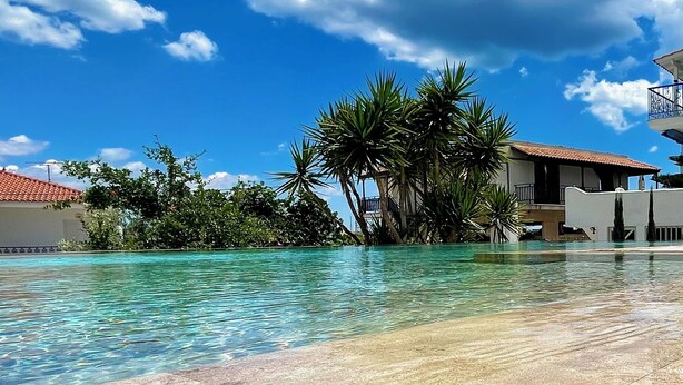 Una piscina cristalina con solárium de baldosas, rodeada de palmeras y un edificio con techo de tejas rojas.