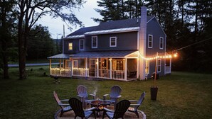 A house with a covered porch, a fire pit with chairs, and string lights. - 4bdrm Cottage central to year round fun w/fireplace (Weston)