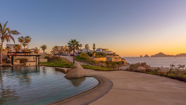 A resort with a swimming pool, palm trees, and a view of the ocean at sunset. - New! Ocean Views, Perfect Location, Great Amenities (Cabo San Lucas)