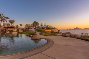 A resort with a swimming pool, palm trees, and a view of the ocean at sunset. - New! Ocean Views, Perfect Location, Great Amenities (Cabo San Lucas)