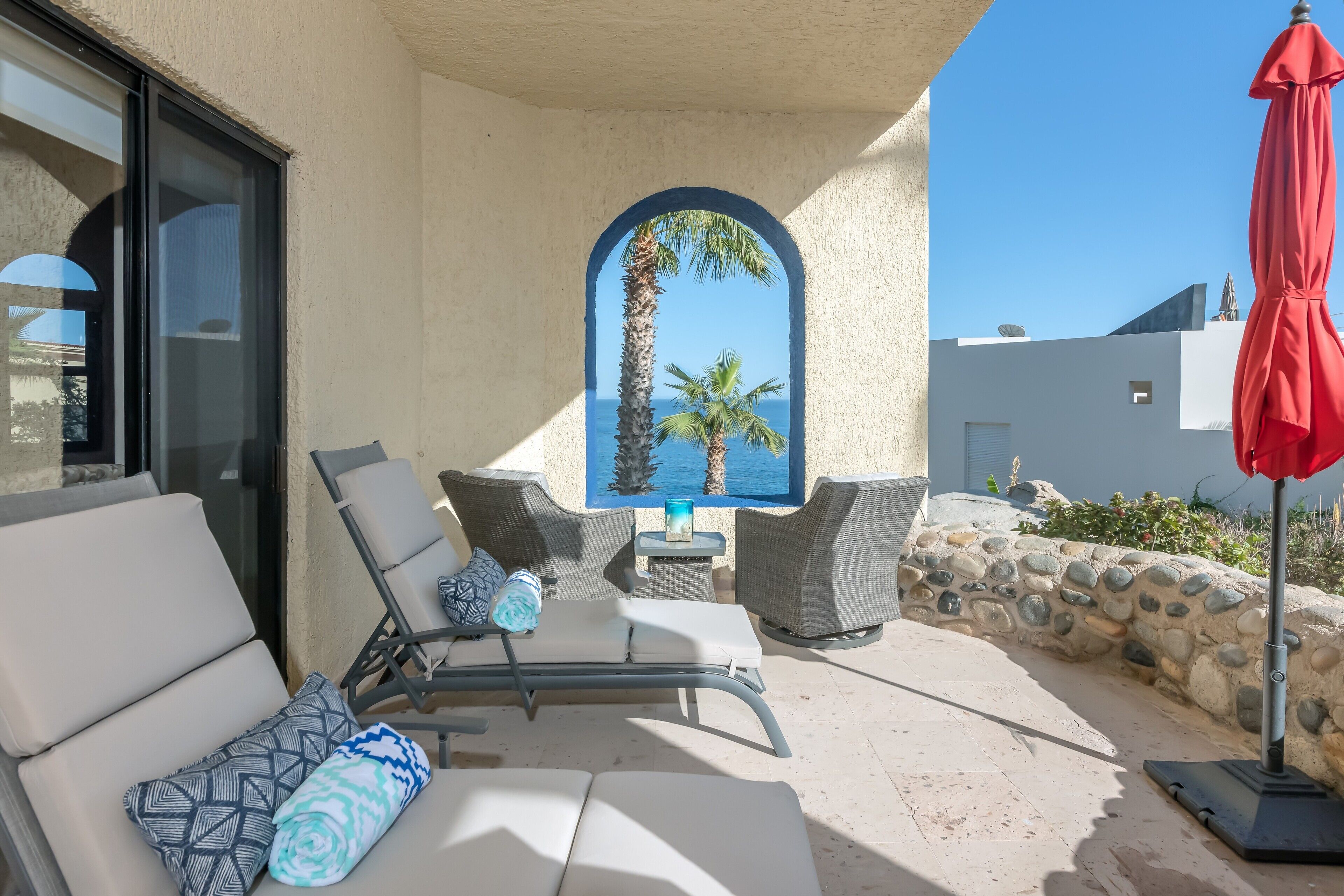 A sunlit outdoor seating area with lounge chairs and a table, overlooking a clear blue sea and palm trees.
