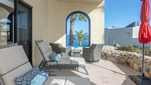 A sunlit outdoor seating area with lounge chairs and a table, overlooking a clear blue sea and palm trees. - New! Ocean Views, Perfect Location, Great Amenities (Cabo San Lucas)