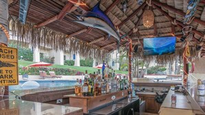 An outdoor bar area with a pool view, bar counter stocked with drinks, and a thatched roof overhead. - New! Ocean Views, Perfect Location, Great Amenities (Cabo San Lucas)