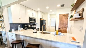 A modern kitchen with white cabinets, a stainless steel sink, a microwave, and a coffee maker. There are two wooden stools and a wooden door. - Adorable 2nd floor Villa with Peek-a-Boo Gulf Views (Panama City Beach)