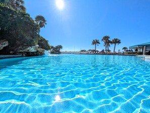 Ein klares, blaues Schwimmbecken mit einer steinigen Klippe und Palmen im Hintergrund.