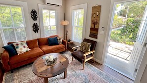 A living room with a brown leather sofa, a wooden coffee table, a wooden chair, and a ceiling fan. There are two windows with blinds, a wall-mounted air conditioner, and a door leading to a balcony. - TOP FLORIDA's La Casita (Fort Myers)